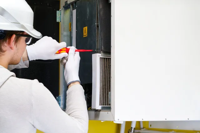 A technician in a white hard hat and gloves uses a screwdriver on equipment.